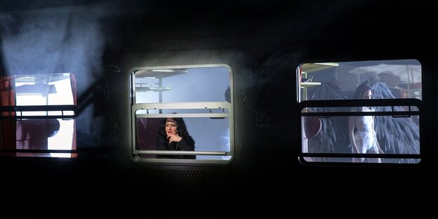 Bühnenbild: Nachtaufnahme auf einen Zug, eine Frau schaut aus dem Fenster, durch ein anderes sehen wir eine Engelsgestalt