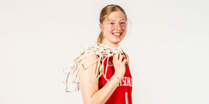Eine junge blonde Frau lächelt in einem roten Trikot die Kamera und hat das Netz eines Basketballkorbes über die Schulter gehängt