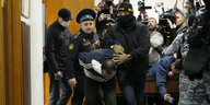 Ein Mann wird von Polzisten in einem Saal geschleppt unter Anwesenheit von vielen Fotografen