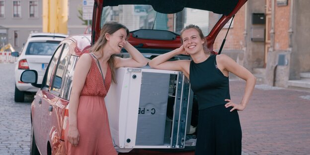 zwei lachende junge Frauen vor dem geöffneten Kofferraum eines Kleinwagens